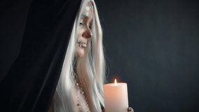 mystical video in Gothic style, woman with blond hair with sweet white skull and black cloak holding burning candle in hands, image for masquerade for Halloween and Day of Dead, lady blows out fire