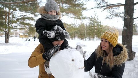 Happy family, mom, son and dad are making a snowman in winter park. Son is sitting on dad's neck