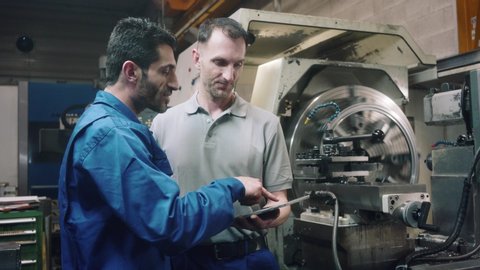 Two workers discussing a project on laptop in front of CNC lathe machine