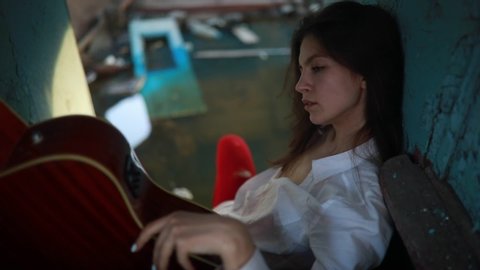 A young sad woman sits with guitar inside of old abandoned ship in white shirt and holey red tights.