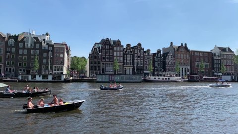 AMSTERDAM - CIRCA JUNE, 2019: Footage of people riding boats on Amstel River in Amsterdam. Historical, old, traditional buildings are in the view. It is a sunny summer day. Camera pans right.