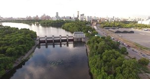 4K cloudy afternoon aerial scenic footage of hydro system dam regulating river water levels overlooking green park and distant large city sky scrapers high rise apartments