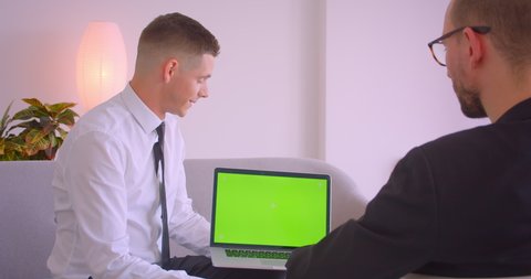 Closeup portrait of young businessman showing laptop with green chroma key screen to partner making offering in the office indoors