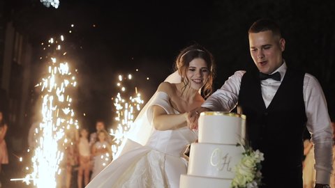 Beautiful young wedding couple eating cake, guests with sparklers. Wedding cake of a gay couple during the wedding party while photographers shooting. Wedding party