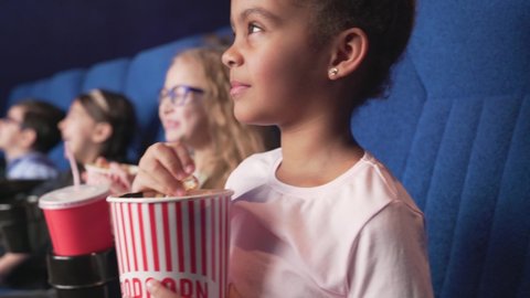 Side view of cheerful little multiracial well dressed children eating tasty popcorn, drinking cola and smiling during funny moments of new comedy. Happy kids spending time together at cinema.