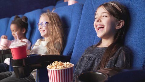 Side view of smiling multinational kids sitting in a row, watching interesting funny cartoon, eating tasty popcorn and spending time at modern cinema. Children look very emotional and happy