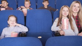 Cheerful little boys and girls watching interesting movie, sitting in comfortable sits and eating tasty popcorn from striped big bucket. Pretty children feeling happy and satisfied at cinema
