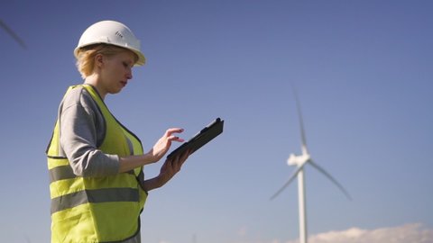 Adult or mature technican woman in special uniform standing with tablet against landscape wind turbine power station on background. Female monitoring system performance and making notes in program
