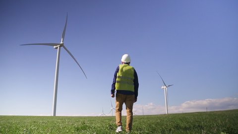 Adult or mature technician man in white hard hat and green uniform wear walking with modern digital tablet wind mill power station on background with blue sky. Male monitoring system performance