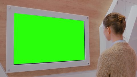 Woman looking at blank digital interactive green display wall at exhibition or museum with futuristic interior. Mock up, future, copyspace, template, chroma key, green screen, technology concept