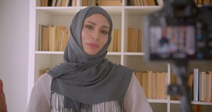 Closeup portrait of young attractive muslim blogger in hijab talking on camera indoors with bookshelves on background