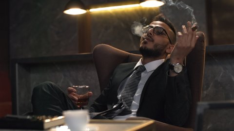 Waist-up shot of mixed race businessman in formal suit, white shirt, tie and glasses relaxing alone after work in private lounge, drinking whiskey, smoking cigar, blowing out smoke and checking watch