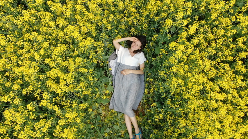 lady in white grey summer dress lies on blooming rapeseed field surrounded by yellow flowers vertical aerial zoom out