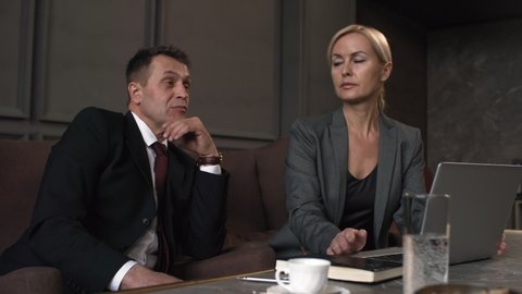Medium shot of Caucasian male executive sitting in armchair and talking with someone during business meeting, and attentive female manager in formal suit looking at him, nodding and typing on laptop