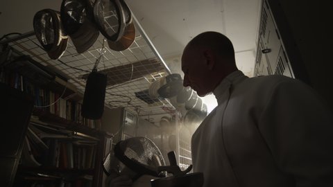 A male fencer puts on his fencing mask in a locker room, lit by a bright window