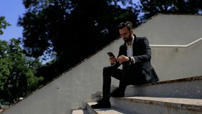 attractive young man businessman with a beard sitting in a park on a concrete staircase and talking on the phone, then looks at the clock on his hand. telephone business negotiations. 4k. 4k video