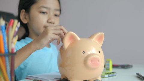Close-up little Asian girl putting coin into the piggy bank metaphor saving money for better future select focus shallow depth of field