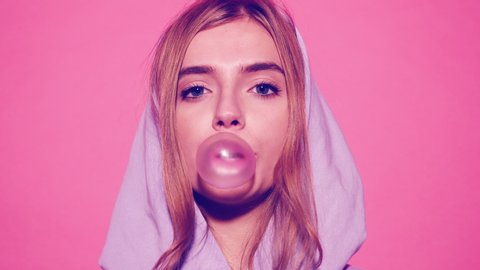 Portrait of young sexy playful blonde girl dressed in blue hoodie standing and isolated over pink background blowing bubblegum candy bubbles and chewing gum. Woman looking at camera. 4k. Slow motion