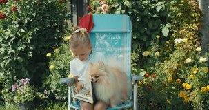Little pretty girl sitting in chair, looking notebook with herbarium. Sunny day, summer house. Interesting hobby. Funny dog playing with kid. 4k raw slow motion footage video 60 fps