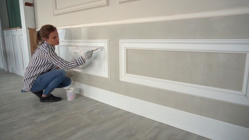 girl in a striped shirt paints a wall with white paint.