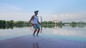 Handsome muscular young man in white shirt, shorts and sports cap exercising with jump rope on lake pier. Young fit man training outdoors during summer days