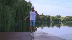 Full length portrait of muscular young man in sport clothing jumping with skipping rope near lake. Confident guy training in the morning time.