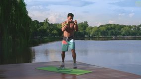 Full length portrait of athletic man with perfect suntanned body training with sport equipments on yoga mat with green nature around. Concept of bodybuilding and and active lifestyle
