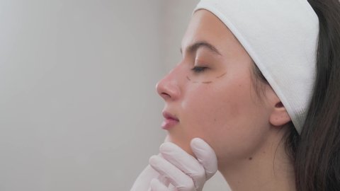Cosmetologist in white rubber gloves touching and drawing correction lines on beautiful woman face in modern clinic. Charming girl with clear skin is getting ready for cosmetology procedures