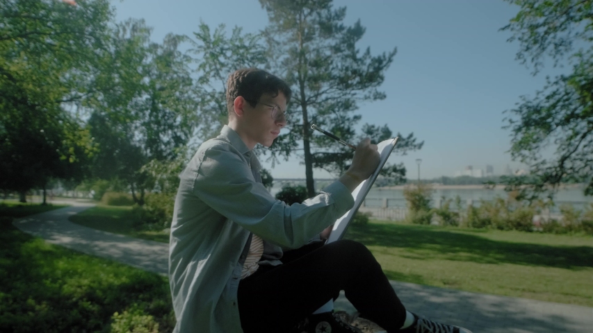 Modern creative young guy in a summer park. He sits on the grass with an album and paints a landscape. The concept of creativity, inspiration, the artist at work. taken from the side.