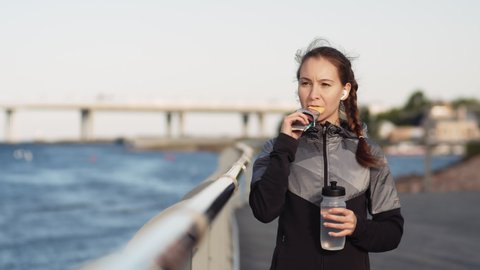 Medium shot of young Caucasian woman standing outdoors on embankment, eating energy bar and holding sport bottle in her hand
