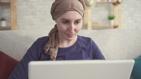positive young woman with cancer in a scarf on her head after chemotherapy uses a laptop