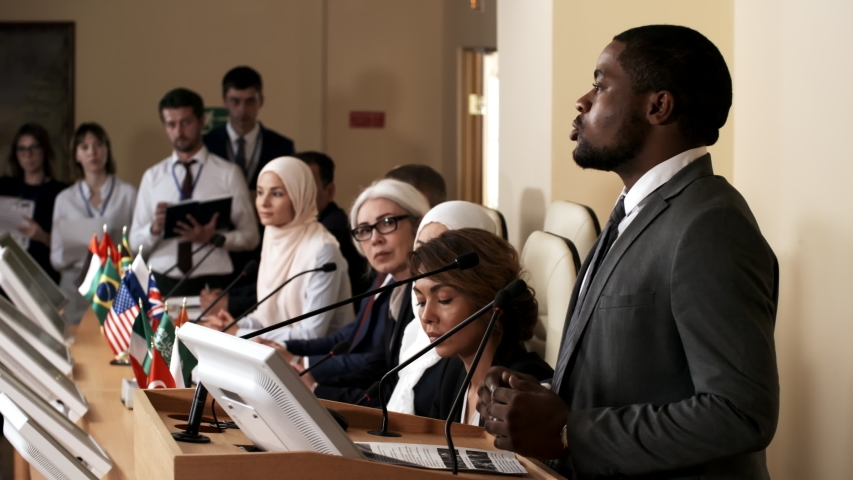 African american man giving a public talk at rostrum during international press conference while multiethnic participants sitting at table with flags of different countries and listening to speech Royalty-Free Stock Footage #1035092261
