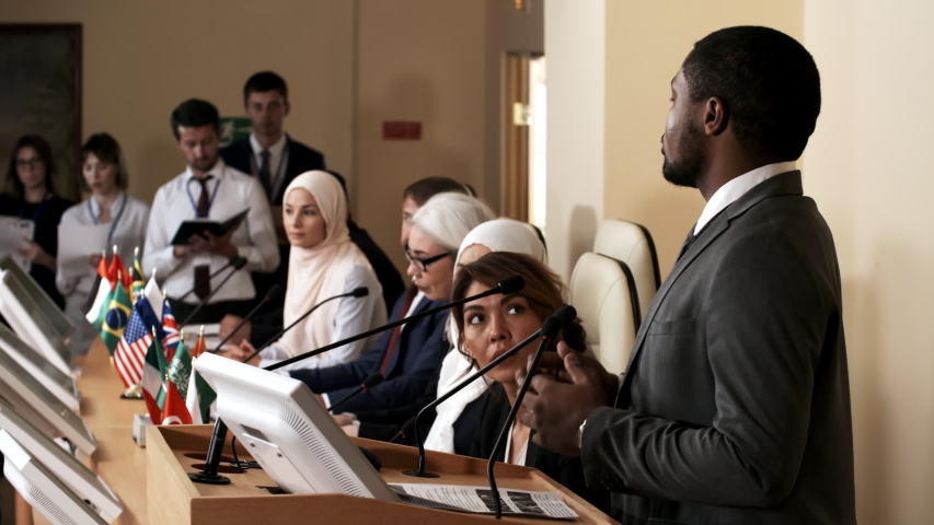 African american man giving a public talk at rostrum during international press conference while multiethnic participants sitting at table with flags of different countries and listening to speech Royalty-Free Stock Footage #1035092261
