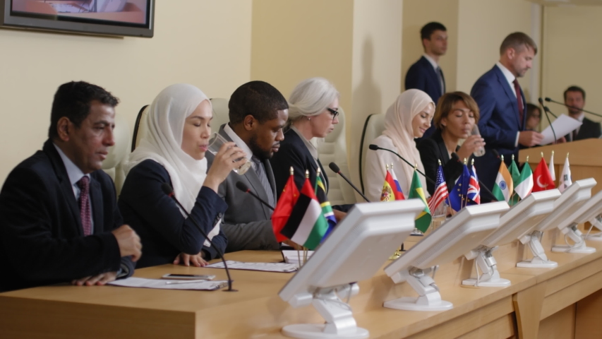 Group of multiethnic male and female politicians walking into conference room, sitting at table with flags and microphones on it, drinking water and talking Royalty-Free Stock Footage #1035104366