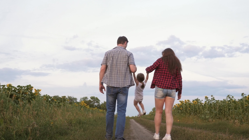 little daughter jumping holding hands mom and dad. Family with small child walks along road and laughs next to field of sunflowers. Mom, dad and daughter are resting together outside city in nature.