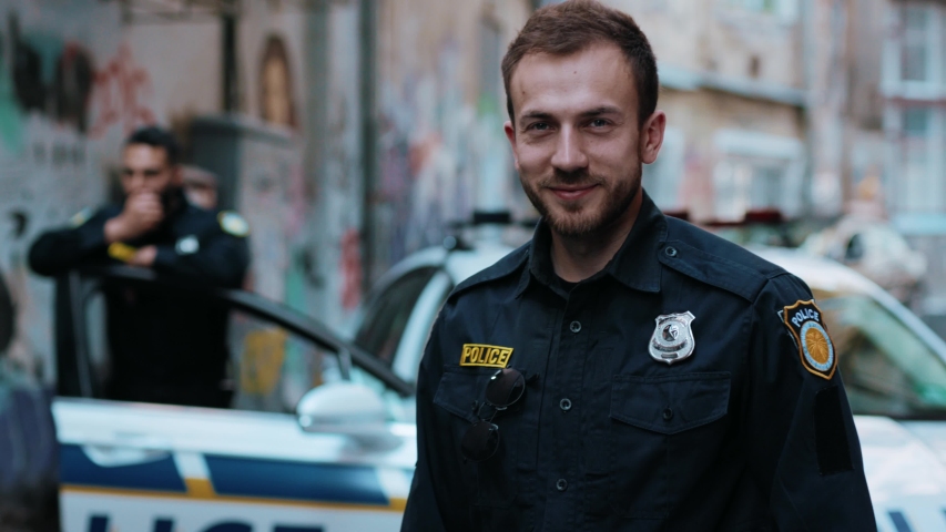 Handsome smiling young man cops stand near patrol car look at camera enforcement officer police uniform auto safety security communication control policeman close up slow motion