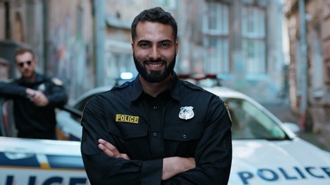 Smiling young man cops stand near patrol car look at camera enforcement happy officer police uniform auto safety security communication control policeman portrait close up slow motion