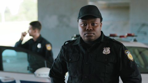 Close up serious face young african american man cops stand near patrol car look at camera enforcement officer police uniform auto safety security communication control policeman close up slow motion