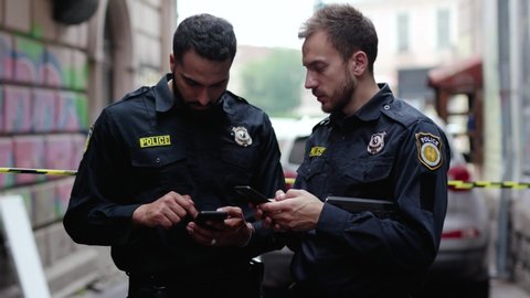 Police at raid. Close-up two handsome policemen searching on smartphones tracking a criminal online. Friendly colleagues cops at police investigation area in the city street.