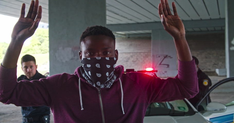 Close-up black man perpetrator wearing mask raising his hands by police order. Police officers on duty arrest an afro-american thief bad guy at daytime. Royalty-Free Stock Footage #1035106649