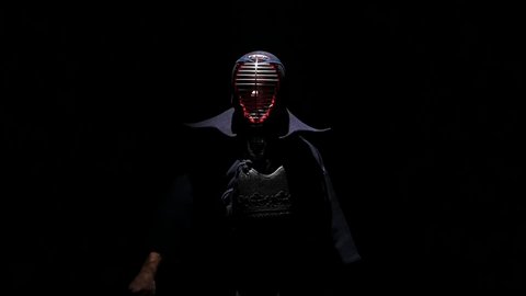 Experienced Kendo warrior wearing in an armor, traditional kimono and helmet, coming out of darkness into the light and looking at the camera. Sportsmanship, slow motion
