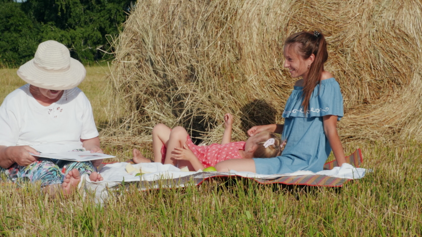 Grandmother, mother and little daughter doing picnic outdoor with a melon in the meadow near the haystack. Positive mood, parenthood, childhood and food concept. Grandmother reading a book to