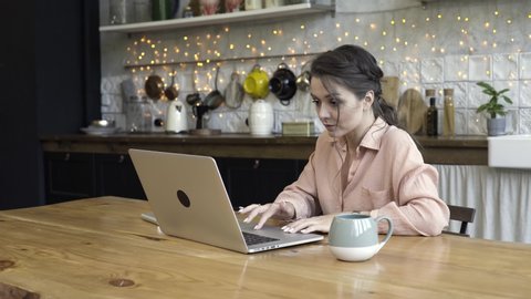 A young woman sitting at the kitchen table using a laptop. with the worried expression on her face. Stock footage. Brunette woman working in her modern designed kitchen.
