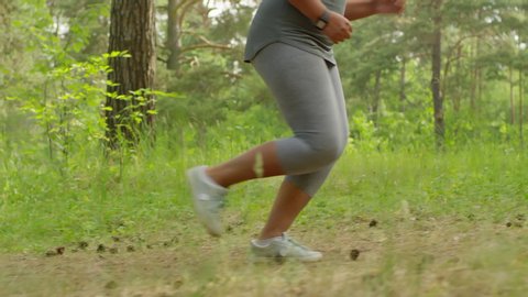 Tracking waist-down shot of unrecognizable fit curvy woman, wearing grey leggings and trainers, running through summer forest with vigorous pace