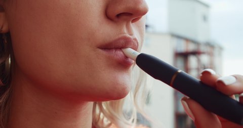 Close-up pretty woman with electronic cigarette outdoors. Young woman smoking tobacco stick heating and exhailing smoke.