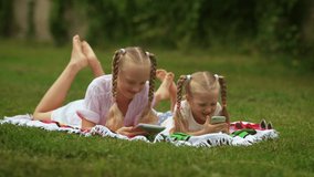 Two kids with gadgets. Two sisters are looking at a display of a tablet and mobile phone, lying on a grass in a backyard in sunny day