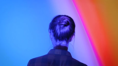 Back view of young woman looking around at modern immersive exhibition or club event with changing multi color projector light illumination. Digital art, technology and entertainment concept
