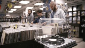 Close-up shot of turntable playing vinyl disc in specialty record shop, and happy diverse customers browsing through merchandise and chatting leisurely in blurred background - Powered by Shutterstock - Get 15% off with code: PIKWIZARD15