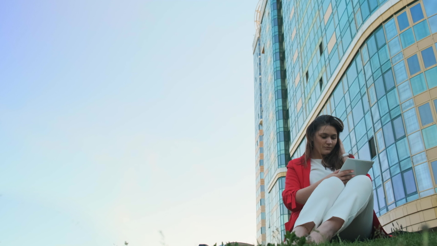 Business woman sitting on the lawn, against the background of a business center surfing in social networks on a tablet