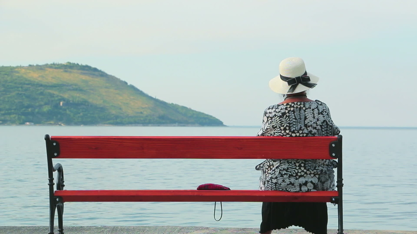 Elderly lady sits alone on the bench and looks at the sea. An old woman sits on a bench by the sea early in the morning